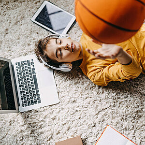 Junge spielt mit deinem Basketball, daneben ist ein Notebook und ein Tablet zu sehen
