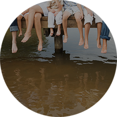 Eltern und Kinder lassen ihre Füße von einem Steg am Wasser herunterhängen.